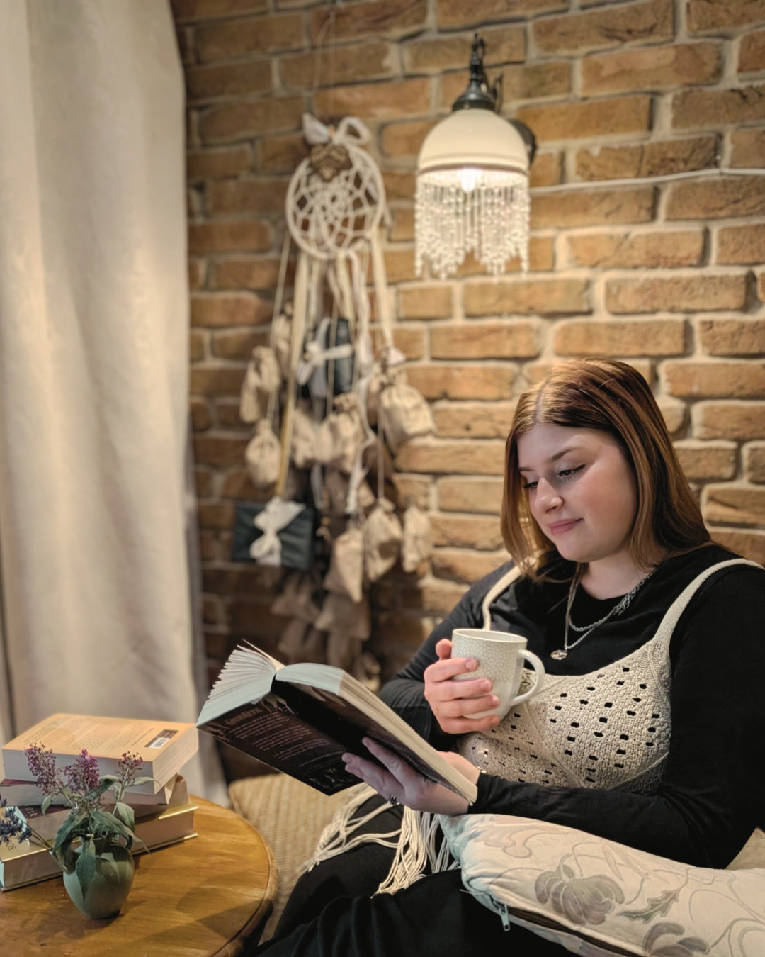 Gemütliche Leseecke mit Hoggynook Adventskalender Dreamcatcher als Weihnachtsdeko an Backsteinwand. Junge Frau entspannt mit Buch und Teetasse im Kerzenschein, warmes Hygge-Ambiente im Dark-Academia- und Boho-Stil. Perfekte Stimmung für Bücherwürmer, Winter- und Weihnachtsgeschenke für Booktok-Community.